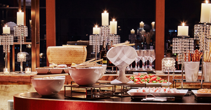 테이블 위에 촛대 장식과 디저트류, 과일이 놓여 있으며, 뒤편으로는 치즈 섹션과 촛대 장식, 창가 야경이 보인다.