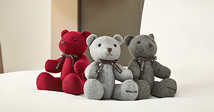 침구위에 신라스테이 베어 3개가 연출되어 있는 이미지. 베어의 목에 리본을 메어져 있고, 왼쪽 발바닥에는 신라스테이 로고가 새겨져 있다.