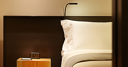 오른편에 베개가 세워진 침대 헤드가 보이고 왼편에는 탁상 위에 시계와 메모패드가 놓여져있다. 