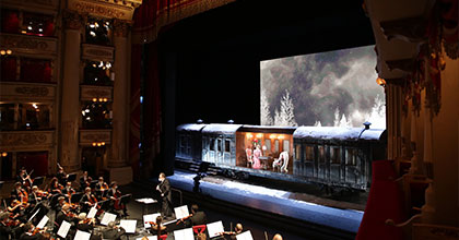 라스칼라 갈라 콘서트의 전경을 보여준다. 왼쪽에는 오케스트라, 오른쪽에는 오페라 무대 위 성악가가 노래를 부르고 있다. 